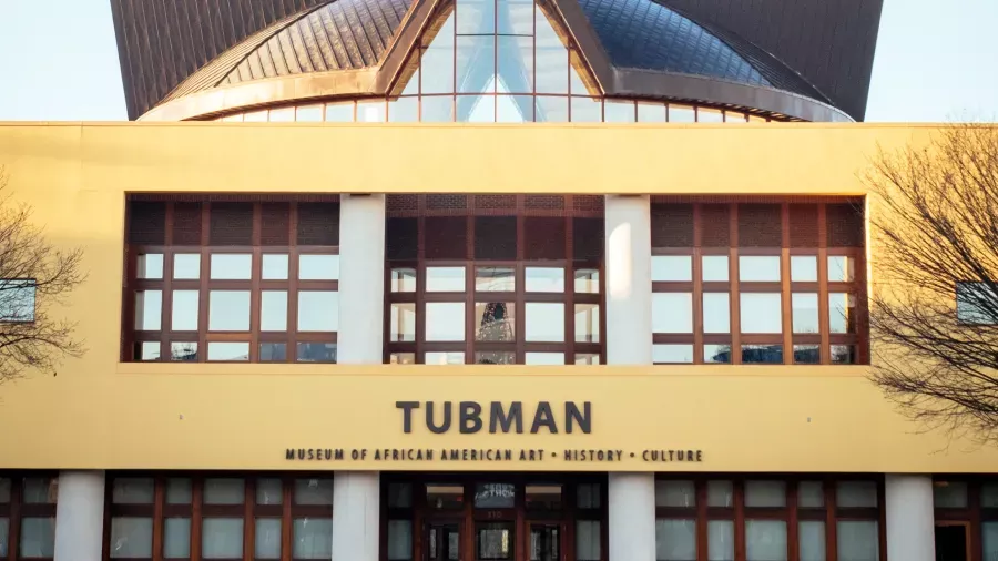 The image shows the exterior of a building labeled "Tubman Museum of African American Art * History * Culture." The building features a yellow facade with large windows and a prominent entrance. The roof has a unique architectural design.