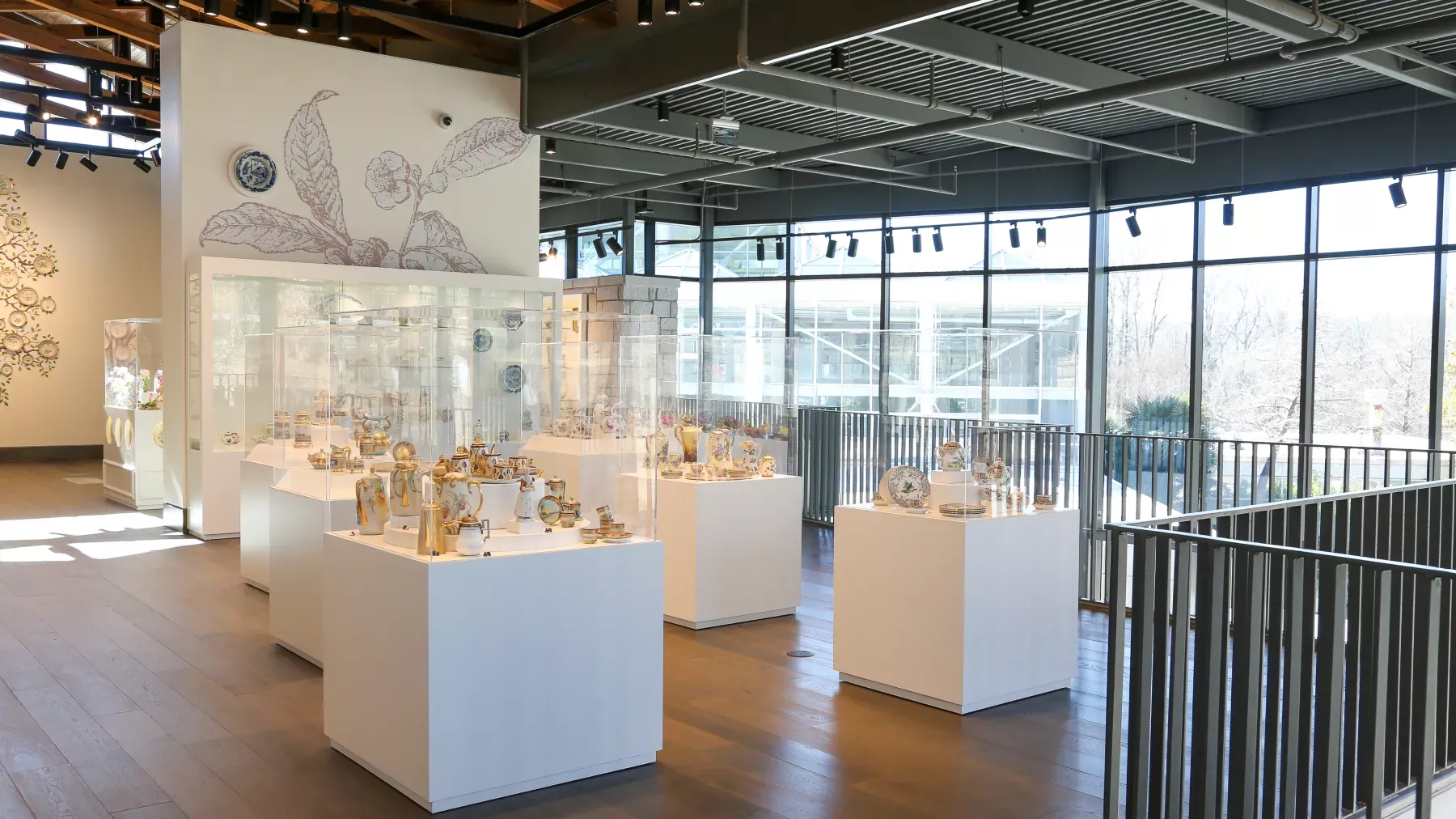 A well-lit museum exhibit featuring various pieces of ceramic art displayed on white pedestals within glass cases. The room has large windows allowing natural light to illuminate the space, wooden floors, and decorative wall art in the background.
