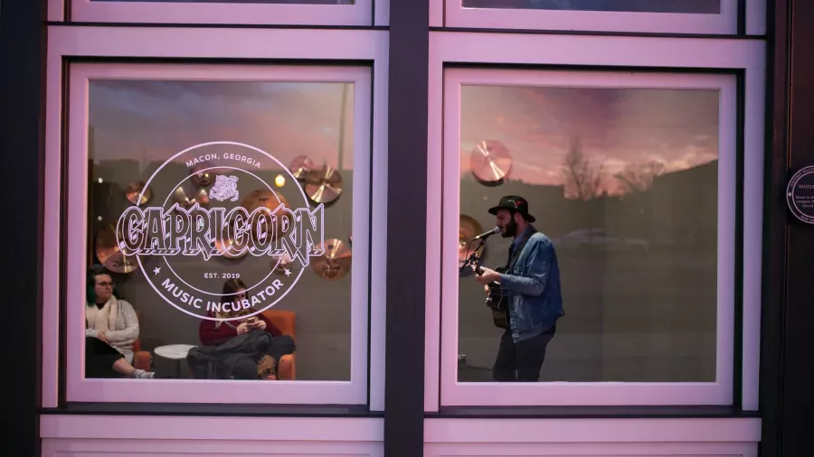 A person playing an acoustic guitar is visible through a large window with a "Capricorn Music Incubator" sign. Inside, other people are seated on a couch listening. The evening sky outside is reflected on the glass, adding a soft glow to the scene.