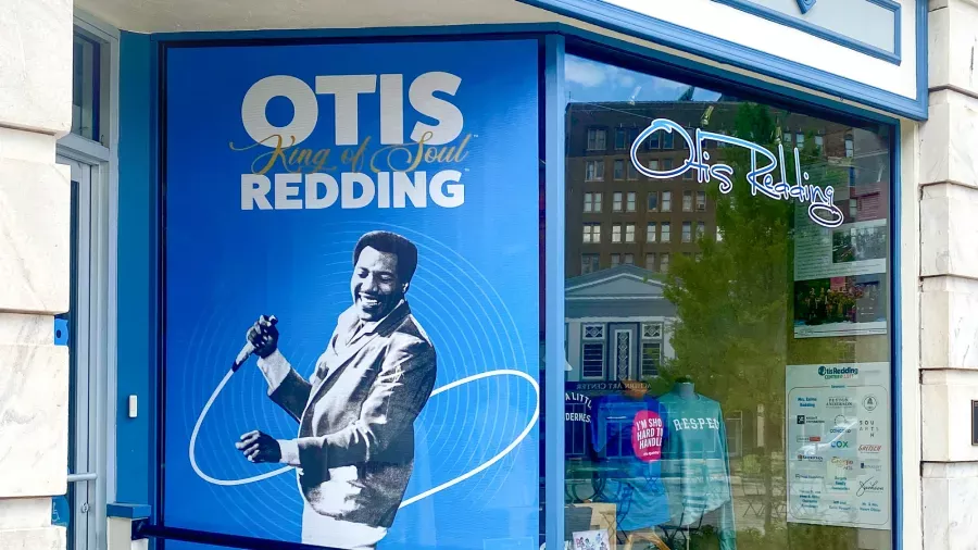 A storefront with a large blue poster of a man holding a microphone. The text on the poster reads "Otis King of Soul Redding." Next to it, a window displays merchandise and "Otis Redding" is written on the glass.