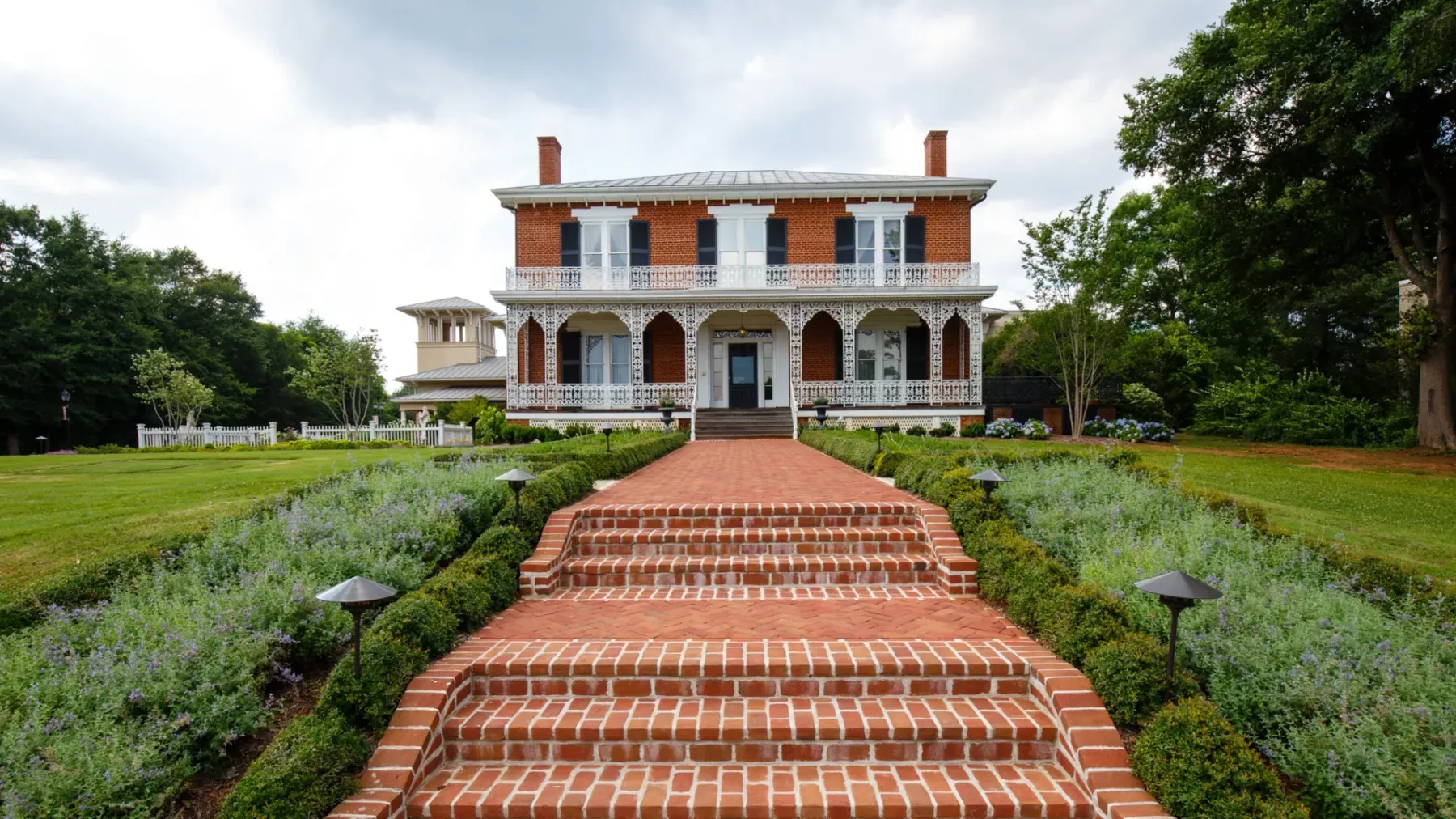 A large, two-story brick house with a wraparound porch and intricate white trim stands at the end of a wide brick path. The pathway features multiple steps leading through well-manicured gardens with low greenery and lantern-style lights. Trees surround the property.
