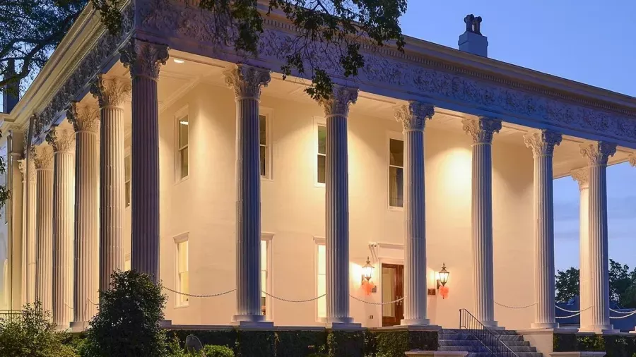 A large, well-lit neoclassical building with tall white columns and a wrap-around porch at dusk. The building's facade is illuminated, highlighting the ornate detailing at the top of the columns. A few windows are visible, and trees frame the structure.