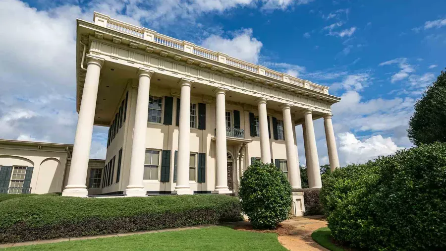 A grand, two-story white mansion with large columns supporting a portico, surrounded by neatly trimmed hedges and lush green bushes. The building stands against a blue sky with scattered clouds, giving it a majestic and historical appearance.