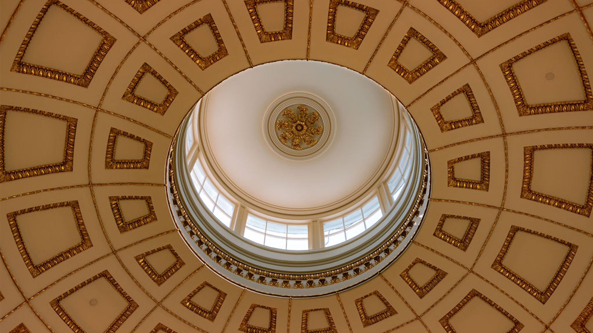 A view from below of an ornately designed circular domed ceiling. The ceiling features concentric circles with decorative gold and white patterns. In the center, there is a skylight ringed by a series of small windows allowing natural light to enter.