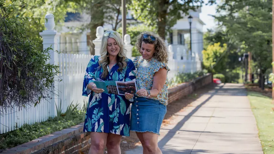 Two women are standing on a sidewalk near a white picket fence, looking at a map or brochure together. One woman is wearing a blue patterned dress, and the other is in a floral blouse and denim skirt. Trees and houses line the background on a sunny day.