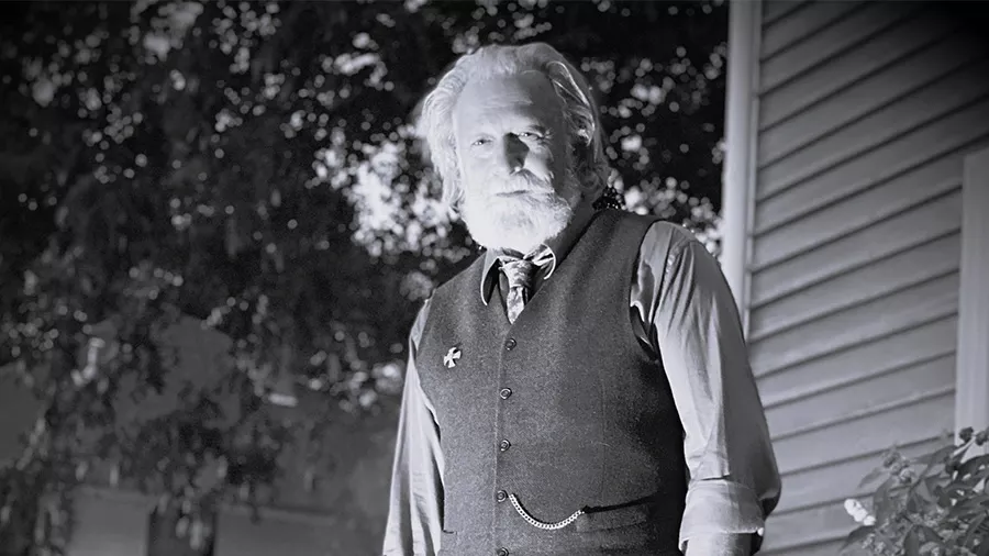 A grayscale photo shows an older man with a long beard and white hair standing outdoors near a building's siding. He is wearing a vest over a shirt and tie, with a pocket watch chain visible. Trees and dark foliage are in the background, evoking the Haunted History often explored in Watkinsville walking tours.