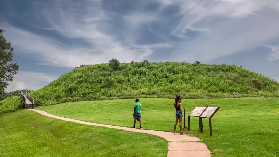 Two visitors stand on a paved path reading informational signs in front of a large, grassy mound. A staircase ascends the mound on the left. The sky is partly cloudy, and the scene is surrounded by greenery.