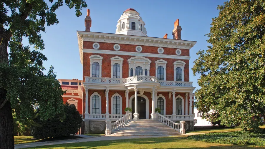 A large, ornate, red-brick mansion with white trim, featuring a grand staircase leading to its entrance. The building has symmetrical windows, a central balcony, and a distinctive, domed cupola on the roof. Lush green trees and a manicured lawn surround the house.