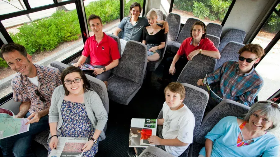 A diverse group of people of various ages are sitting on a bus. Some are holding magazines and books, engaging in reading. The bus is filled with gray seats and has large windows, allowing greenery outside to be visible. Everyone appears relaxed and content.
