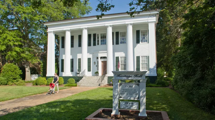 A white, two-story, classical-style building named "Heritage Hall" features tall columns and a landscaped front yard. A woman pushes a red stroller along the pathway leading to the entrance. A large sign in the foreground provides information about the hall.