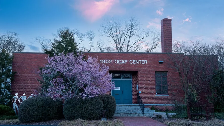 The image shows a brick building labeled "1902 OCAF CENTER" at twilight. In the foreground, a blooming tree with pink flowers and manicured bushes adds charm. The Oconee Cultural Arts Foundation's building has a chimney, a central door with steps leading up to it, and a serene sky overhead.