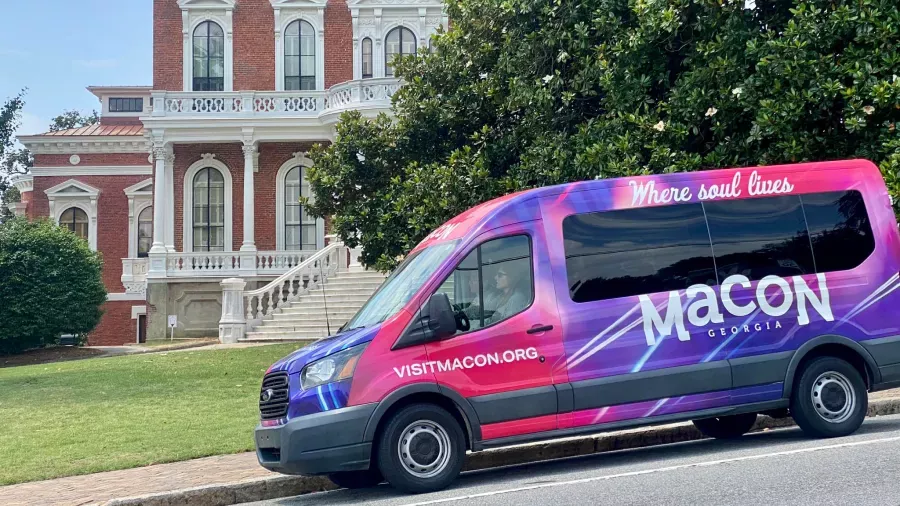 A colorful van with "Macon Georgia" and "Where soul lives" inscribed on its side is parked in front of a historic red brick building with white columns and a grand staircase. The van also has the website address "visitmacon.org" displayed.