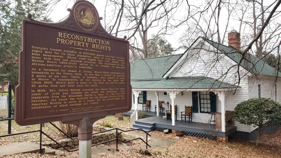 A historical marker titled "Reconstruction Property Rights" stands in front of a white house with a green roof, surrounded by trees. The marker details the history of property ownership and reconstruction in Georgia, particularly related to African-American ownership.