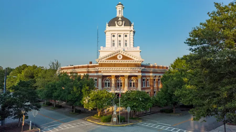 A historic courthouse building with a prominent clock tower and dome, surrounded by trees and situated at the intersection of two streets. The structure features columns and intricate architectural details, with a clear blue sky in the background.