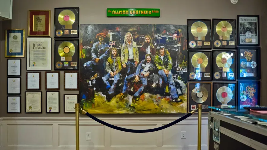 The image shows a wall display with a large painting of a music band, surrounded by framed awards, certifications, and numerous gold and platinum records. The display is roped off with a velvet rope barrier. A green sign at the top reads "The Allman Brothers Band.