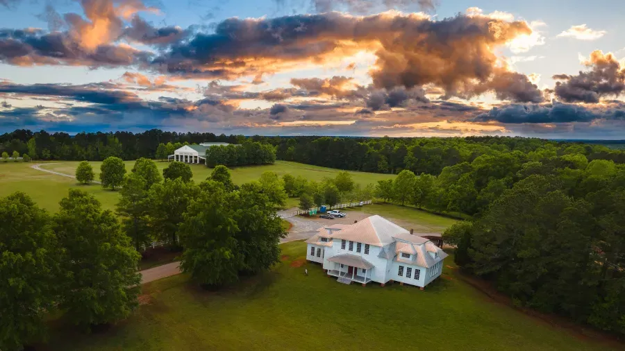 Aerial view of a large white house surrounded by green trees and fields at sunset. A second building in the distance sits amidst the expansive landscape. The sky is filled with dramatic clouds tinged with orange and pink hues from the setting sun.