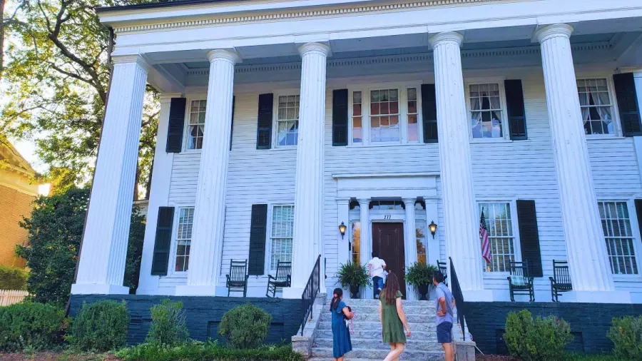 People gathered in front of a large white two-story colonial-style house with tall columns and black shutters. Some stand near the entrance while others walk on the front walkway. The house is surrounded by greenery and trees in the background.