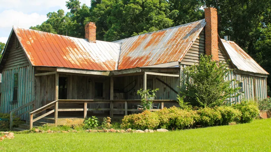 A rustic, weathered wooden house with a corrugated metal roof stands amid green grass and trees. The roof shows signs of rust and wear, and two brick chimneys are visible. A wooden porch runs along the front, partially covered by a roof extension.