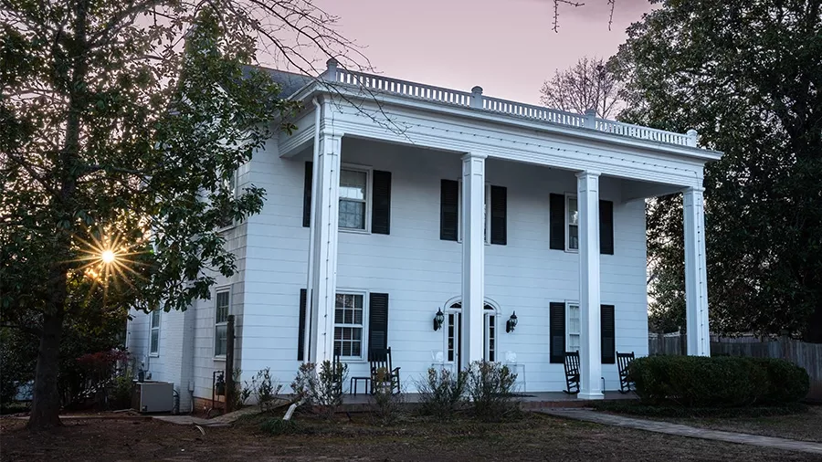 A stately white two-story house, reminiscent of the Jackson House, with four tall columns supporting an extended portico. The house features black shutters and a symmetrical design. Two rocking chairs sit on the front porch as the sun sets behind trees, casting a warm glow over the scene.