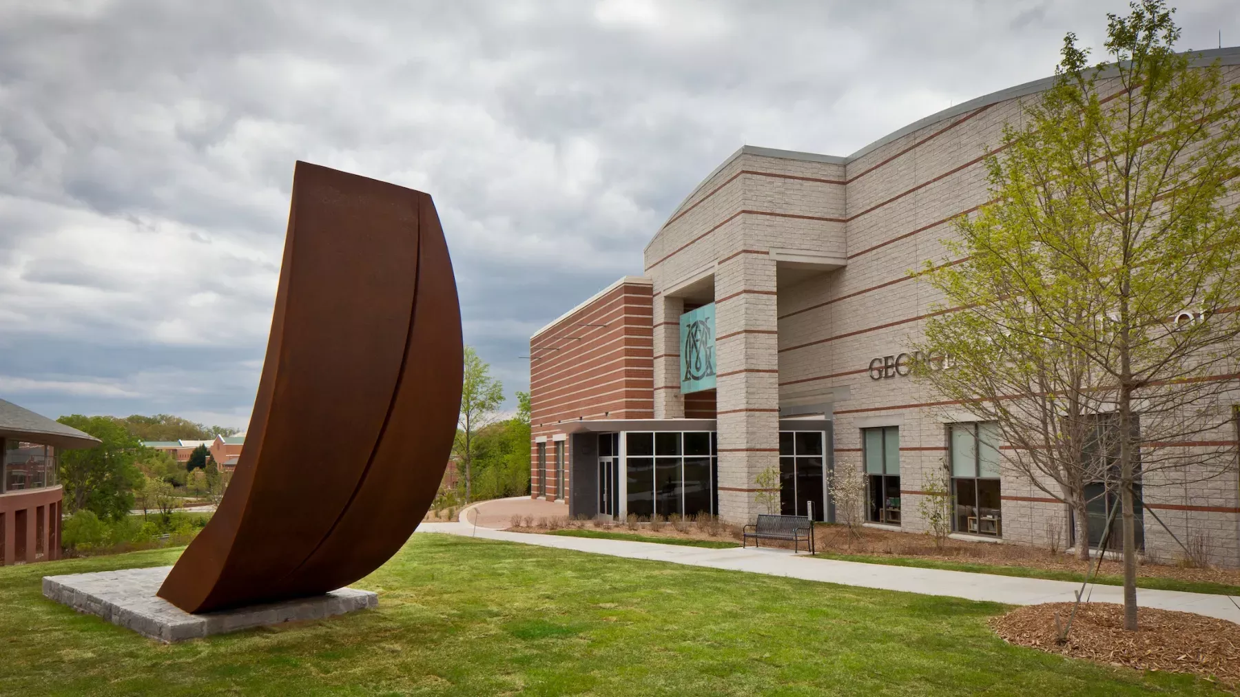 A modern building with a brick and stone facade. In front, there is a large, curved metal sculpture on a stone base. Nearby, there are trees with budding green leaves. The sky is overcast with gray clouds.