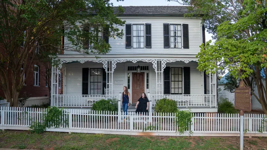 Two people stand in front of a historic wooden house with a white picket fence, intricate porch detailing, and dark shutters on the windows. Trees frame both sides of the house, and a large commemorative plaque is visible to the right.