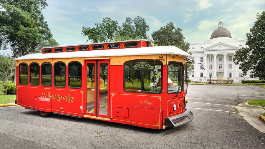 A vibrant red trolley with "Milledgeville" written on its side is parked in front of a large white building with columns and a dome. Trees surround the area, and the sky is partly cloudy. The trolley is named "Milly.