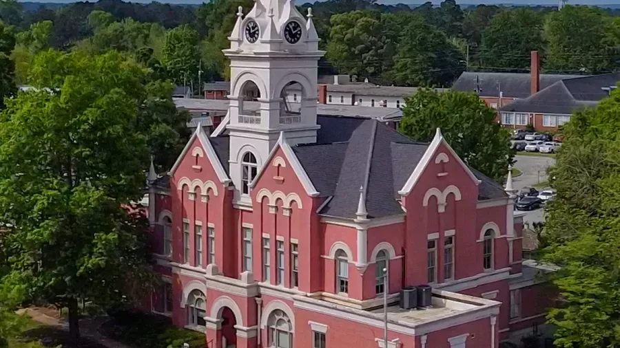 Aerial view of a historic red brick building with white accents and a clock tower. The building is surrounded by green trees and has a backdrop of a small town with a few buildings and a parking lot visible.