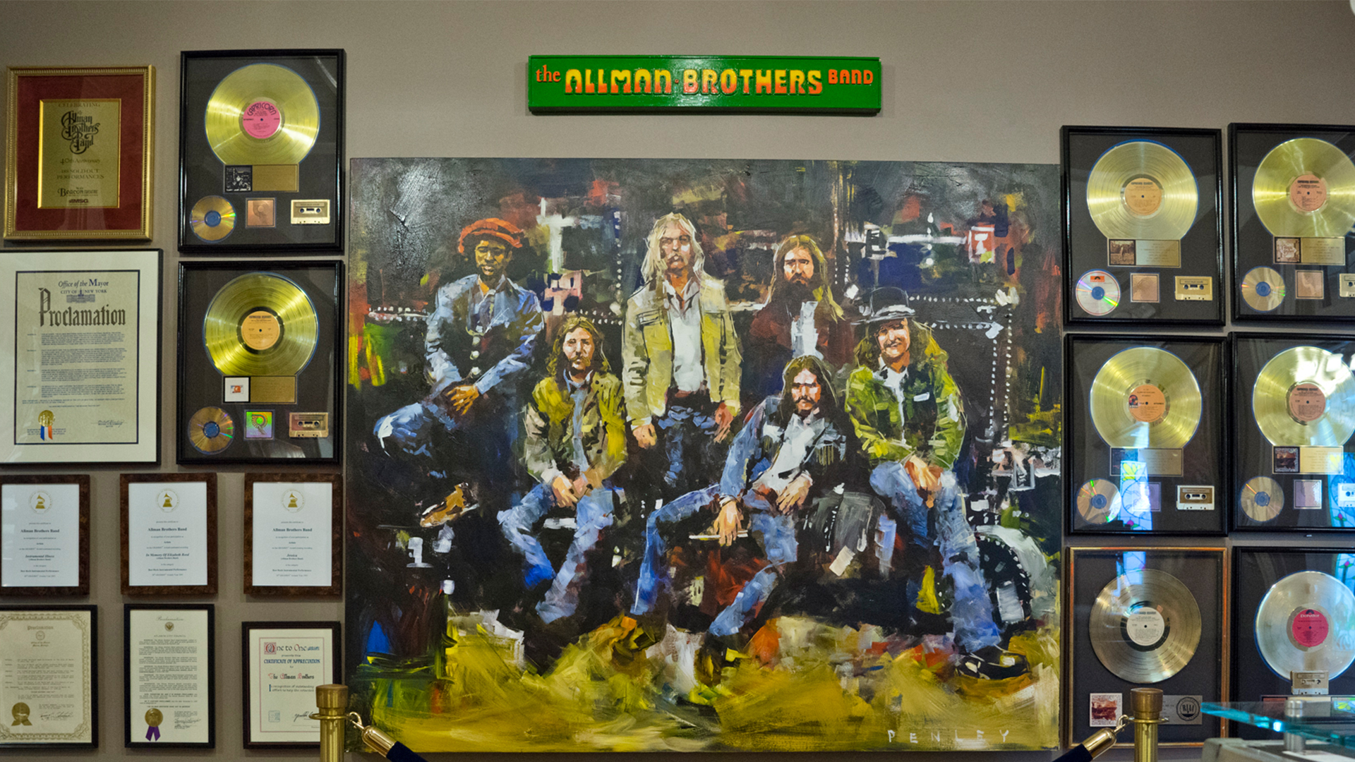 A wall display featuring a large painting of the Icons of American Music, surrounded by numerous framed gold and platinum records, plaques, and certificates. Above the painting is a green sign with yellow text that says "The Allman Brothers Band.