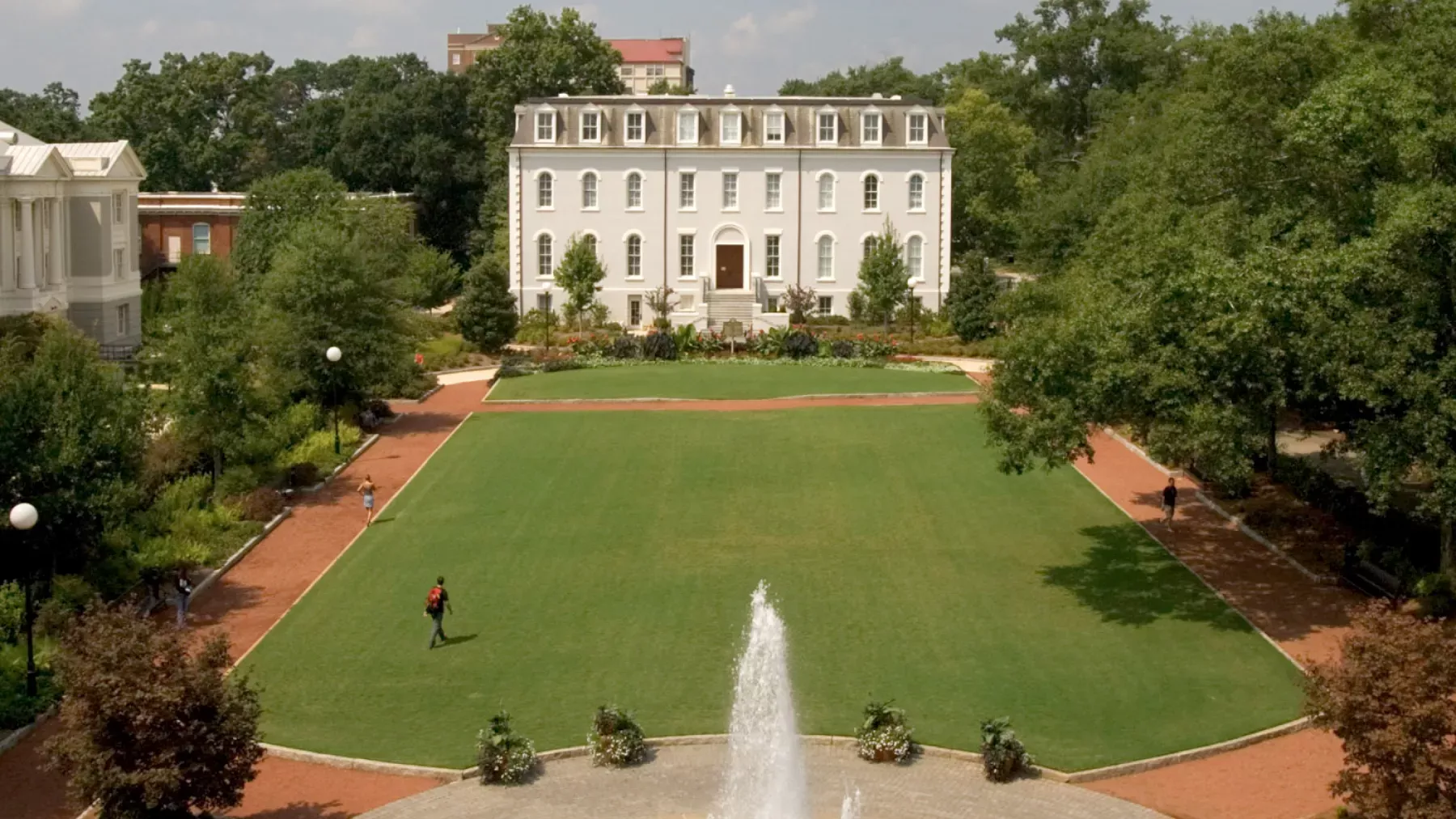 Aerial view of a university campus showing a large, well-manicured lawn bordered by brick pathways. In the background is a stately, white three-story building with multiple windows. A central fountain is in the foreground, and a few people walk along the paths.