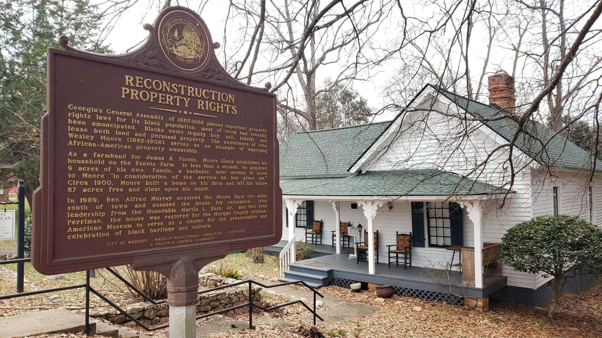 A historical marker titled "Reconstruction Property Rights" stands in front of an old white wooden house with a green roof. The marker describes land ownership by African-American families post-1865 in Georgia, with the house partially visible behind trees.