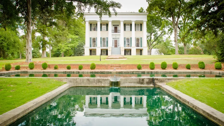A stately white mansion with tall pillars stands amidst lush greenery. In front, a rectangular reflective pool mirrors the mansion's facade. Trimmed hedges and trees line the spacious lawn. The setting is serene and picturesque.