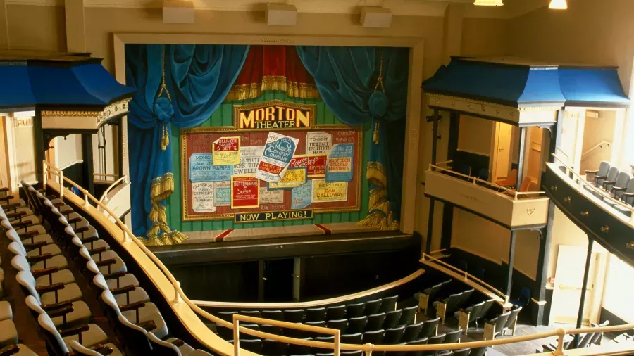 A theater with a grand stage featuring a colorful backdrop reading "Morton Theater" adorned with various posters. The seating area includes rows of cushioned chairs, balconies on either side, and elegant architectural details throughout.