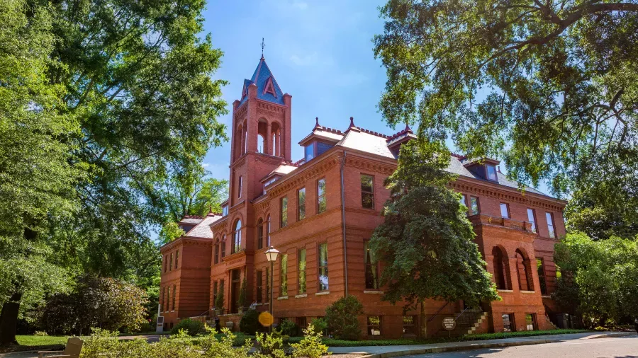 A historic, multi-story brick building with a prominent tower stands amidst lush green trees under a clear blue sky. The building features tall, arched windows and intricate architectural details, conveying a sense of classic elegance and charm.