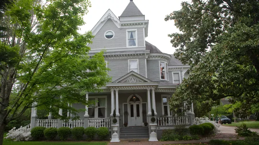 A large, two-story Victorian-style house with a prominent turret and wrap-around porch. The house is painted a light gray with white trim, and it is surrounded by lush green trees and shrubs. The architecture includes intricate details and columns on the porch.