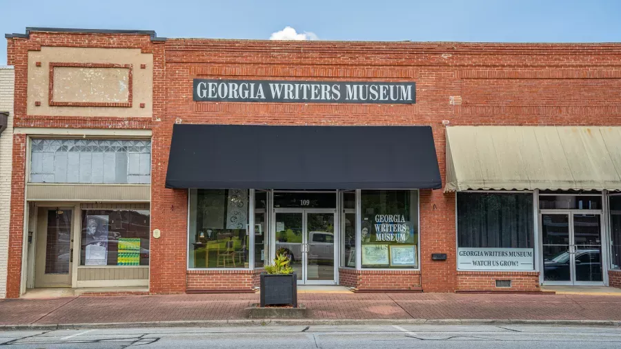A red brick building with a black awning houses the Georgia Writers Museum. The entrance features large glass doors and windows. The sign "Georgia Writers Museum" is prominently displayed above the awning. The neighboring storefronts have beige and green awnings.