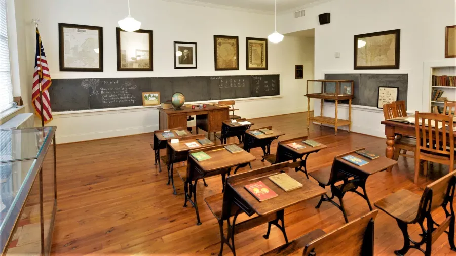 A vintage classroom with wooden desks arranged in rows, each desk holding a book. A teacher's desk with a globe and books is at the front. Blackboard writing is visible, with framed maps and pictures on the walls. An American flag stands near the windows.