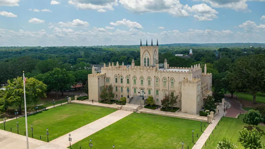 Aerial view of an elegant, historic, castle-like building with pointed towers, surrounded by lush green lawns and trees. The clear sky above is dotted with fluffy white clouds. Pathways lead to the building, and an American flag stands in the foreground.