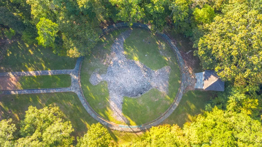 Aerial view of the Great Serpent Mound, a prehistoric effigy mound in the shape of a serpent, located in a grassy clearing surrounded by dense trees. A stone path leads to the mound, and a small shelter stands nearby.