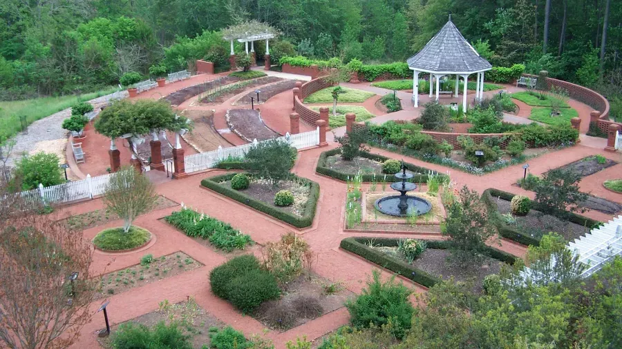 Aerial view of a formal garden with brick pathways and organized plant beds, featuring a central fountain, a white gazebo, tiered seating area, and various trees and shrubs. The garden is surrounded by dense greenery and forest.