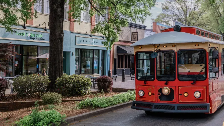 A red trolley car is moving down a street lined with shops and outdoor seating. Trees provide shade over the scene and people sit at tables outside the "Local Well Cafe." The setting appears to be a charming, small-town or suburban area.