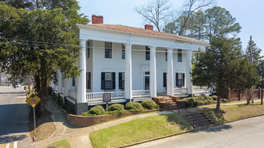Large, two-story white building with a red metal roof and multiple tall columns at the entrance. It features a wraparound porch, black shutters, a historical marker, and is surrounded by trees and greenery at the intersection of two streets.