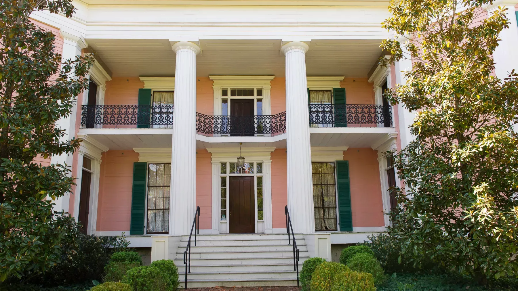 A two-story, pink historic building with large white columns, green shutters, and intricate black wrought-iron balcony railings. The entrance features a dark wood door, flanked by windows. The front garden has neatly trimmed bushes, and two tall trees frame the building.