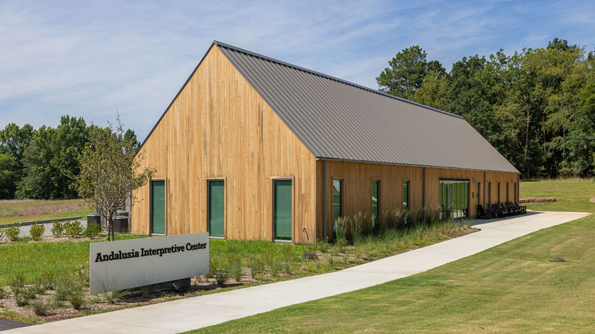 A modern wooden building with a metal roof and several green doors sits under a clear sky. A paved pathway leads to the entrance, flanked by grasses and small shrubs. A sign in front reads "Andalusia Interpretive Center." Trees are in the background.