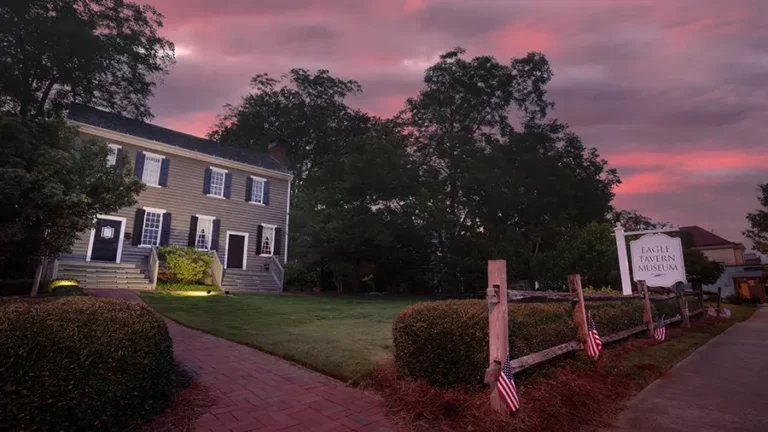 A historic building stands under a dramatic pink and purple sky at sunset. American flags adorn the fence in front. A sign reads "Eagle Tavern Museum." Well-kept shrubs and a brick pathway lead up to the entrance of Eagle Tavern.