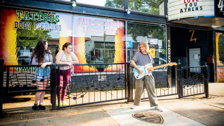A person with long hair is playing an electric guitar on a sidewalk in front of a tattoo and piercing studio. Two people stand behind a metal fence, watching and engaging with the guitarist. The studio has colorful signage and a marquee above.