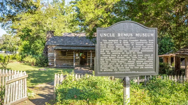 A sign in front of the Uncle Remus Museum reads: "Uncle Remus Museum: This memorial to Joel Chandler Harris, born in Eatonton Dec. 8, 1848, was constructed from three slave cabins found in Putnam County." The museum and log cabin structures are visible in the background.