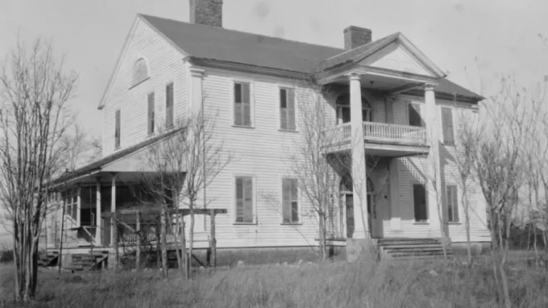 A black and white photo of a large, two-story wooden house with a prominent front porch supported by columns. The house features multiple windows and a chimney. Leafless trees and overgrown grass surround the property.