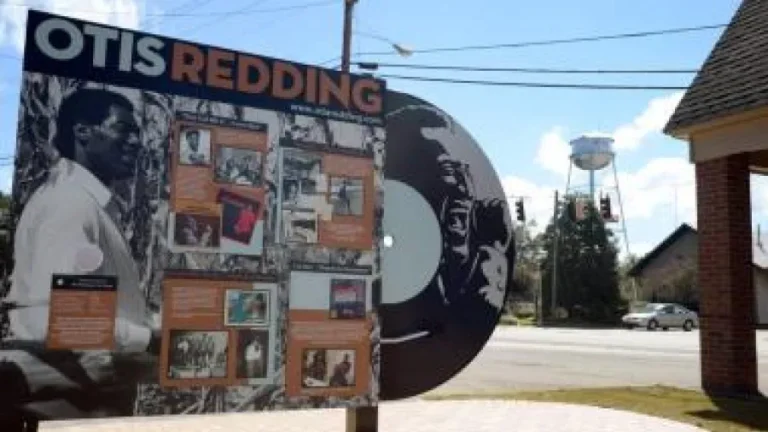 A display board and large vinyl record art dedicated to Otis Redding are situated outdoors near a street. The sign features photos and information about the musician. A water tower and buildings are visible in the background under a partly cloudy sky.