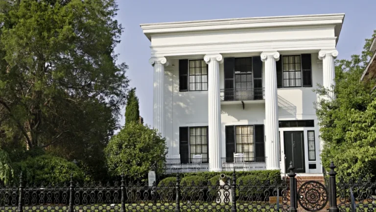 A two-story white house with four large columns in the front, featuring a black front door and shutters. The house is surrounded by lush greenery and a decorative black wrought-iron fence. A tall tree stands to the left side of the house.