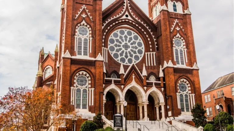 A large, historic red-brick church with intricate architectural details, including two tall towers and a prominent circular stained glass window above the main entrance. The entrance has three arched doorways, and a staircase with railings leads up to it.